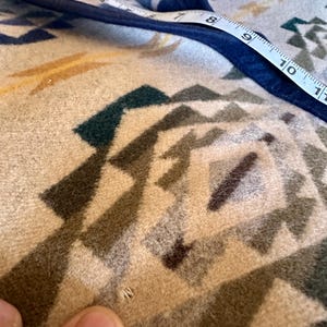 Może przedstawiać: Zbliżenie beżowo-niebieskiego koca z geometrycznymi wzorami. Miarka pokazuje krawędź koca. Koc ma odcienie beżu, brązu, zieleni i niebieskiego. Krawędź koca jest obszyta ciemnoniebieską lam&oacute;wką.