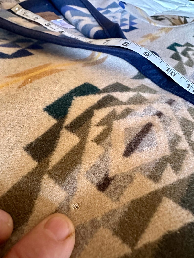 Może przedstawiać: Zbliżenie beżowo-niebieskiego koca z geometrycznymi wzorami. Miarka pokazuje krawędź koca. Koc ma odcienie beżu, brązu, zieleni i niebieskiego. Krawędź koca jest obszyta ciemnoniebieską lam&oacute;wką.