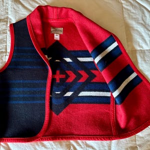 May include: A sleeveless vest with a red collar and trim. The vest features a geometric pattern in red, white, and blue on a black background. The interior is red with a geometric design. A small tag is visible.