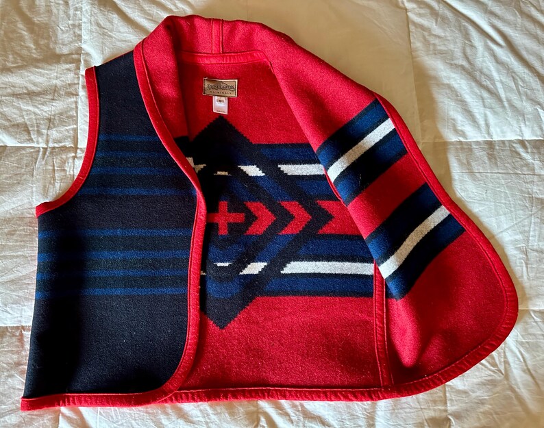 May include: A sleeveless vest with a red collar and trim. The vest features a geometric pattern in red, white, and blue on a black background. The interior is red with a geometric design. A small tag is visible.