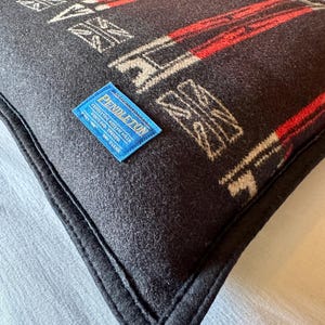 Peut inclure: Un coussin carr&eacute; gris fonc&eacute; avec un motif g&eacute;om&eacute;trique rouge et blanc cass&eacute;. Le coussin a une bordure noire et une &eacute;tiquette bleue avec le mot "Pendleton". Le coussin est fait d'une mati&egrave;re tiss&eacute;e douce.