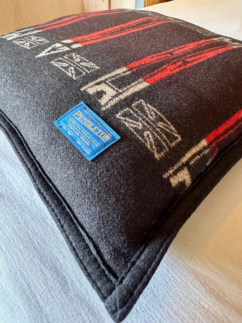 Peut inclure: Un coussin carr&eacute; gris fonc&eacute; avec un motif g&eacute;om&eacute;trique rouge et blanc cass&eacute;. Le coussin a une bordure noire et une &eacute;tiquette bleue avec le mot "Pendleton". Le coussin est fait d'une mati&egrave;re tiss&eacute;e douce.