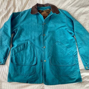 Può includere: Una giacca in tela blu turchese con colletto in pelle marrone e diverse tasche. La giacca ha una chiusura con bottoni e un'etichetta marrone con il testo "Woolrich" sul petto sinistro.