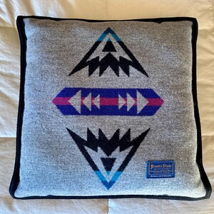 Op de afbeelding: Grijs vierkant kussen met een zwarte rand en geometrische patronen in zwart, blauw en roze. Het kussen heeft een label met de tekst "Beaver State" en "Robes and Shawls."
