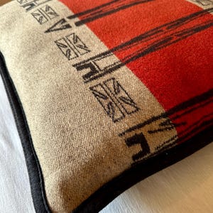 Peut inclure: Coussin carr&eacute; avec un motif &agrave; rayures rouges et noires sur fond beige. Le coussin a une bordure noire et un motif g&eacute;om&eacute;trique dans les coins. Le tissu est doux et textur&eacute;.