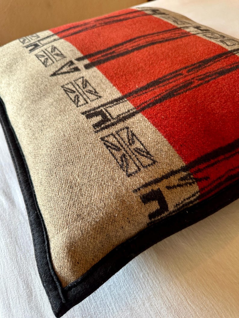 Peut inclure: Coussin carr&eacute; avec un motif &agrave; rayures rouges et noires sur fond beige. Le coussin a une bordure noire et un motif g&eacute;om&eacute;trique dans les coins. Le tissu est doux et textur&eacute;.