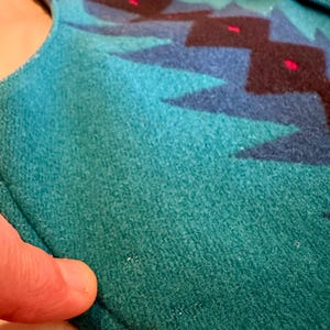 May include: Close-up of a teal and blue patterned fabric. The design features geometric shapes in shades of blue, black, and red. The fabric appears to be a soft, textured material, possibly wool or a similar textile. A silver button is visible.