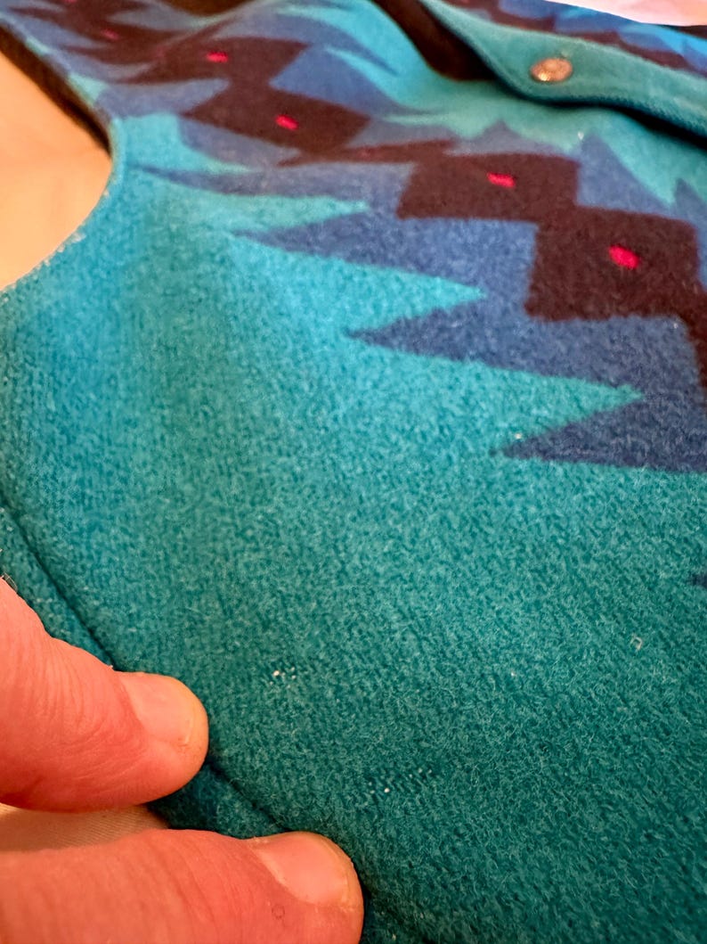 May include: Close-up of a teal and blue patterned fabric. The design features geometric shapes in shades of blue, black, and red. The fabric appears to be a soft, textured material, possibly wool or a similar textile. A silver button is visible.