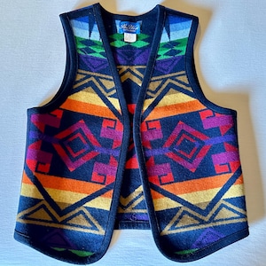 May include: A colorful, open-front vest with a geometric pattern in shades of orange, red, purple, yellow, and blue. The vest has a navy blue background and black trim. The label reads "Rio Vista".