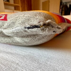 May include: Close-up of a grey pillow with a red and white design on one side. The pillow has a striped pattern in shades of orange, yellow, and purple on the other side. The pillow is resting on a light grey surface.