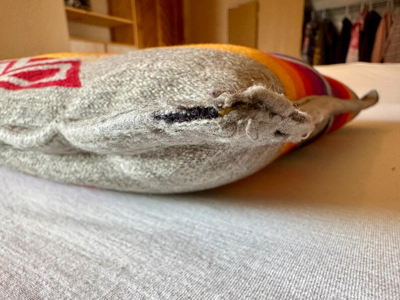 May include: Close-up of a grey pillow with a red and white design on one side. The pillow has a striped pattern in shades of orange, yellow, and purple on the other side. The pillow is resting on a light grey surface.