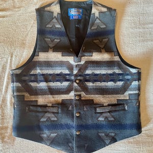 Peut inclure: Un gilet Pendleton arborant un motif géométrique dans des tons gris anthracite, bleu marine et beige. Le vêtement présente une encolure en V, une doublure intérieure noire, quatre boutons métalliques sur le devant et deux poches passepoilées.