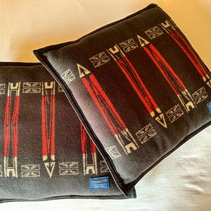 Peut inclure: Deux coussins carr&eacute;s avec un fond gris fonc&eacute; et un motif de rayures verticales rouges et de formes g&eacute;om&eacute;triques blanches. Les coussins ont un passepoil noir et une petite &eacute;tiquette bleue avec du texte blanc.