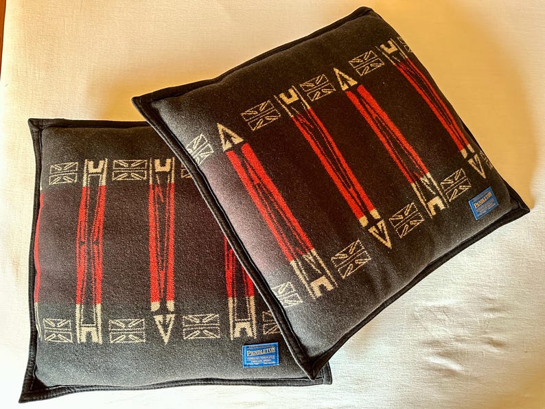 Peut inclure: Deux coussins carr&eacute;s avec un fond gris fonc&eacute; et un motif de rayures verticales rouges et de formes g&eacute;om&eacute;triques blanches. Les coussins ont un passepoil noir et une petite &eacute;tiquette bleue avec du texte blanc.