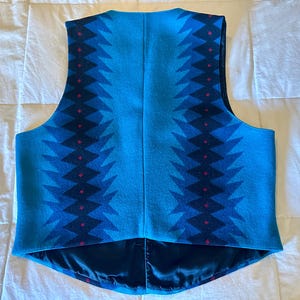 May include: A turquoise vest with a geometric pattern in shades of blue and red. The vest has a satin lining and a classic cut, suitable for layering. The design features a repeating zig-zag pattern.