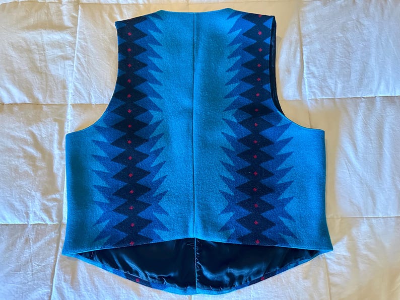 May include: A turquoise vest with a geometric pattern in shades of blue and red. The vest has a satin lining and a classic cut, suitable for layering. The design features a repeating zig-zag pattern.