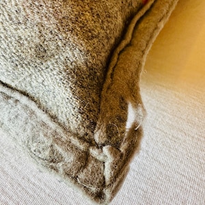 May include: Close-up of a grey and orange cushion. The cushion has a textured, woven appearance with a visible seam. The corner of the cushion is visible, showing the fabric and stitching details. The cushion is likely made of wool or a similar material.