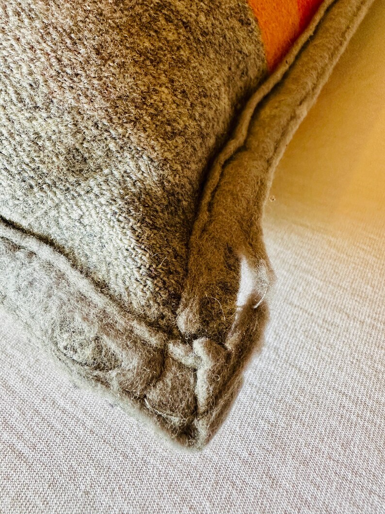 May include: Close-up of a grey and orange cushion. The cushion has a textured, woven appearance with a visible seam. The corner of the cushion is visible, showing the fabric and stitching details. The cushion is likely made of wool or a similar material.
