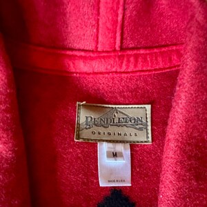May include: Close-up of a red Pendleton Originals jacket. The jacket has a hood and a label with the brand name and size M. The jacket is made in the USA.
