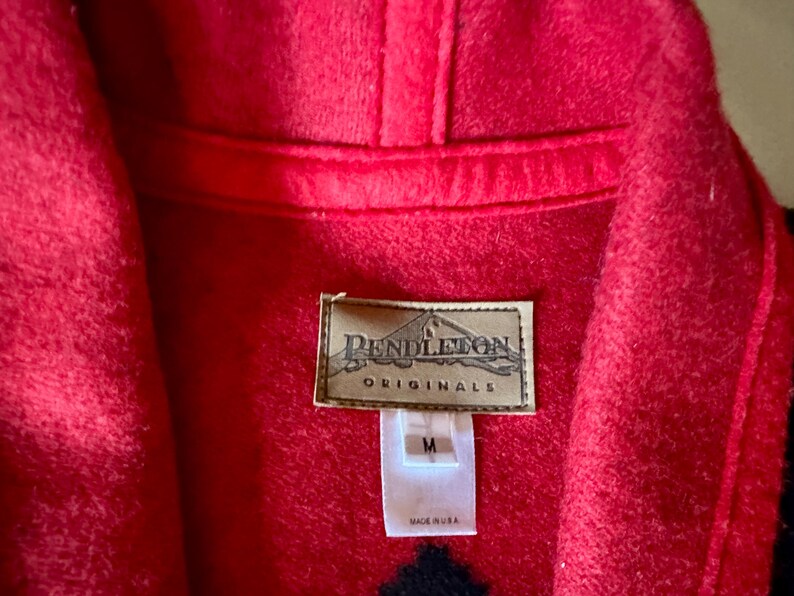 May include: Close-up of a red Pendleton Originals jacket. The jacket has a hood and a label with the brand name and size M. The jacket is made in the USA.