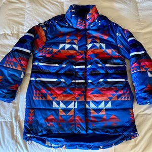 May include: A blue puffer jacket with a geometric pattern in shades of red, white, and black. The jacket has a high collar and a zip closure.