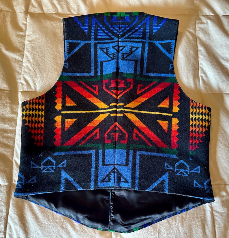 Puede incluir: Un chaleco negro con un vibrante patr&oacute;n geom&eacute;trico en azul, rojo, naranja, amarillo y verde. El chaleco tiene un forro negro y un dise&ntilde;o de cuello en V. El patr&oacute;n presenta formas angulares y dise&ntilde;os sim&eacute;tricos.