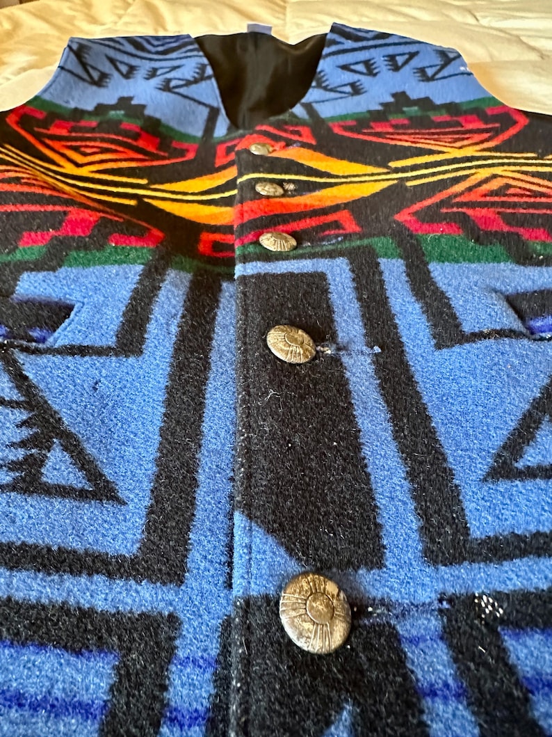 Puede incluir: Un chaleco de lana con un patr&oacute;n geom&eacute;trico del suroeste en azul, negro, rojo, amarillo y verde. El chaleco tiene un forro negro y botones plateados. El chaleco es de talla mediana.