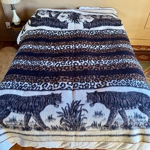 May include: A decorative blanket featuring a tiger and leopard print design. The blanket has a cream-colored base with stripes of leopard print, dark blue, and brown. Two tigers are depicted on the bottom edge.