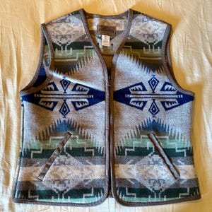 May include: A zip-up vest featuring a geometric pattern in shades of gray, blue, green, and white. The garment has a brown trim along the edges and two front pockets. A Pendleton label is visible at the inner neckline with a white size tag.