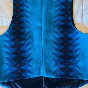 May include: A turquoise vest with a geometric pattern in shades of blue and black, accented with small red diamonds. The vest has a black satin lining and a V-neck design. The back of the vest is visible.