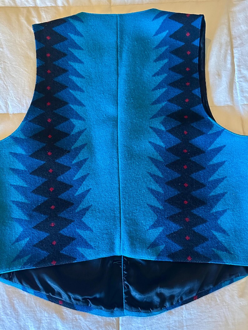 May include: A turquoise vest with a geometric pattern in shades of blue and black, accented with small red diamonds. The vest has a black satin lining and a V-neck design. The back of the vest is visible.