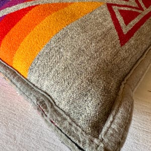 May include: A grey cushion with a geometric design. The design features a rainbow of colours, including red, orange, and yellow, with red and purple geometric shapes. The cushion has a grey border.