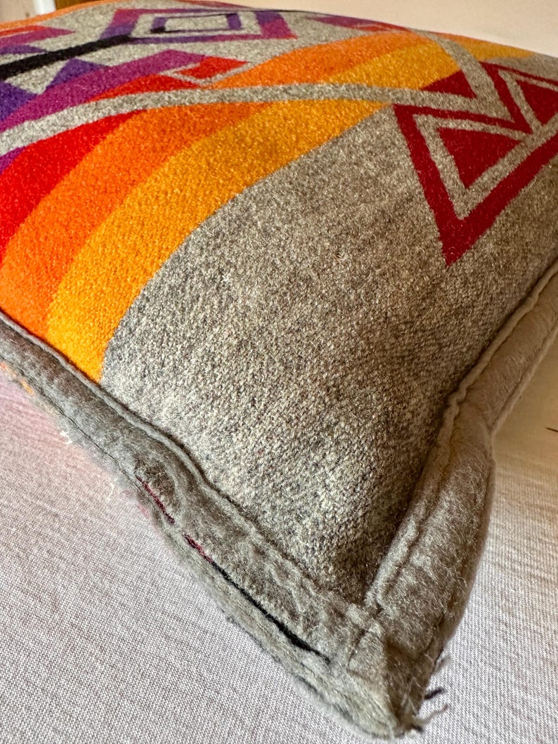 May include: A grey cushion with a geometric design. The design features a rainbow of colours, including red, orange, and yellow, with red and purple geometric shapes. The cushion has a grey border.