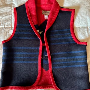 May include: A sleeveless vest with a red collar and trim. The vest features a black body with blue horizontal stripes and a red and white patterned design on the front. The vest is made of a thick, woven material.
