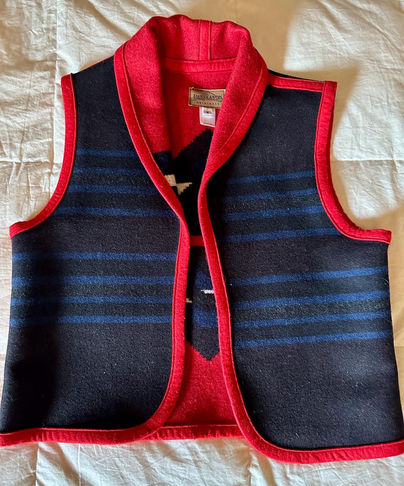 May include: A sleeveless vest with a red collar and trim. The vest features a black body with blue horizontal stripes and a red and white patterned design on the front. The vest is made of a thick, woven material.