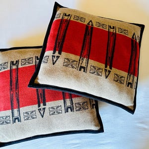 Peut inclure: Deux coussins carr&eacute;s avec un fond beige, des rayures rouges et des motifs g&eacute;om&eacute;triques noirs. Les coussins ont des bordures noires. Le design est sym&eacute;trique et les coussins sont plac&eacute;s sur une surface blanche.