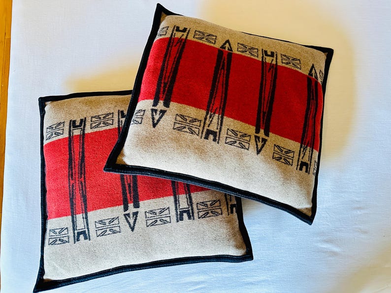 Peut inclure: Deux coussins carr&eacute;s avec un fond beige, des rayures rouges et des motifs g&eacute;om&eacute;triques noirs. Les coussins ont des bordures noires. Le design est sym&eacute;trique et les coussins sont plac&eacute;s sur une surface blanche.