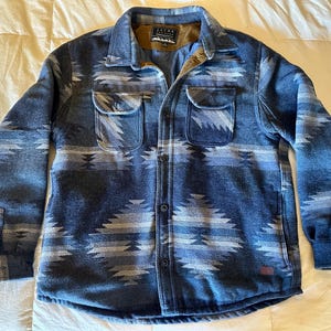 Könnte beinhalten: Ein blaues Hemd mit Knöpfen und einem Muster im Südwest-Stil in Blau- und Grautönen. Das Hemd hat zwei Brusttaschen und einen Kragen. Der Stoff scheint ein mittelschweres Material zu sein, das für kühleres Wetter geeignet ist.