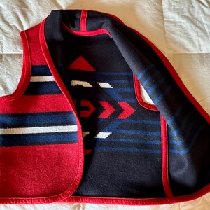 May include: A red, white, and blue patterned vest with a geometric design. The vest has a red trim and a black interior with red, white, and blue geometric patterns. The vest is made of a thick, woven material.