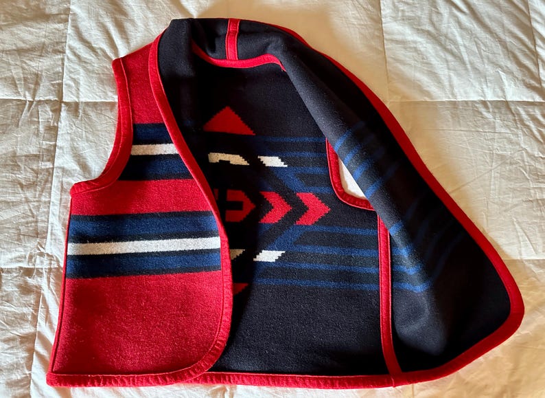 May include: A red, white, and blue patterned vest with a geometric design. The vest has a red trim and a black interior with red, white, and blue geometric patterns. The vest is made of a thick, woven material.