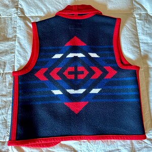 May include: A navy blue vest with a geometric pattern in red, white, and blue. The vest has a red collar and trim. The back of the vest features a diamond-shaped design with a cross in the center.