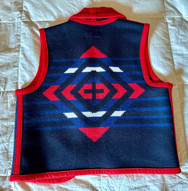 May include: A navy blue vest with a geometric pattern in red, white, and blue. The vest has a red collar and trim. The back of the vest features a diamond-shaped design with a cross in the center.