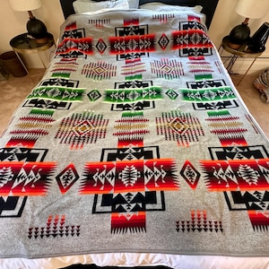 May include: A large rectangular blanket featuring a grey base with a repeating geometric pattern in vibrant shades of red, orange, yellow, green, and black. The design includes symmetrical diamond and cross motifs arranged in a grid across the surface.
