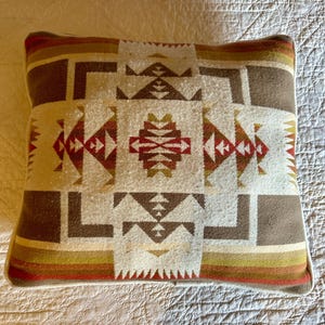 Puede incluir: Cojín rectangular con un patrón geométrico en tonos marrón, rojo, crema y verde. El cojín tiene un borde blanco y una apariencia texturizada, lo que sugiere una tela tejida. El diseño presenta formas y líneas angulares.