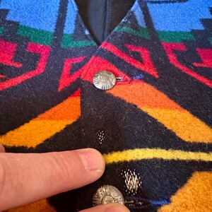 Puede incluir: Primer plano de un chaleco colorido con un patr&oacute;n geom&eacute;trico en azul, rojo, verde, naranja y amarillo. El chaleco tiene botones plateados. La tela parece ser lana gruesa o similar.