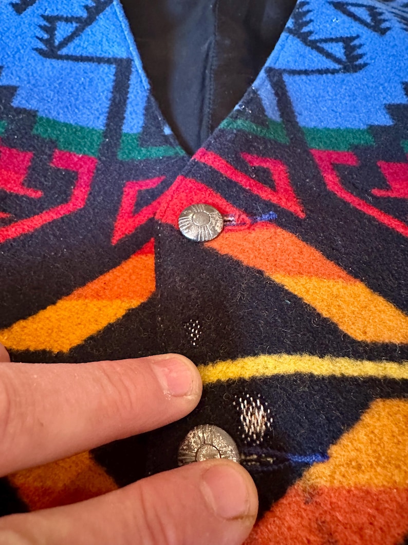 Puede incluir: Primer plano de un chaleco colorido con un patr&oacute;n geom&eacute;trico en azul, rojo, verde, naranja y amarillo. El chaleco tiene botones plateados. La tela parece ser lana gruesa o similar.