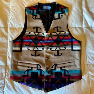 May include: A tan vest with a geometric pattern in black, red, blue, and white. The vest has a V-neck, a black lining, and four buttons down the front. The vest has two front pockets and a label on the back.