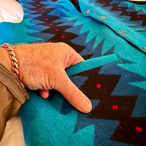 May include: A turquoise and blue patterned vest with a black lining and gold buttons. The vest features a geometric design with red accents. The pocket is open, revealing the interior lining. The vest is made of a thick, woven material.