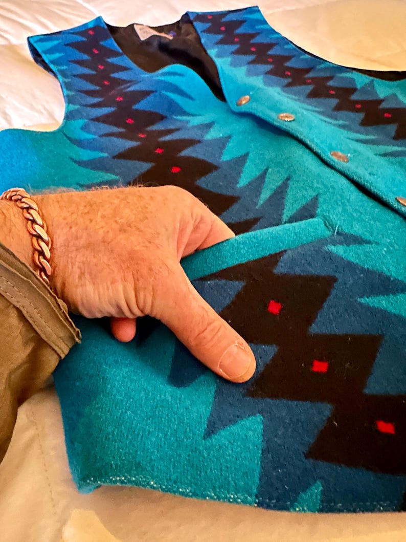 May include: A turquoise and blue patterned vest with a black lining and gold buttons. The vest features a geometric design with red accents. The pocket is open, revealing the interior lining. The vest is made of a thick, woven material.