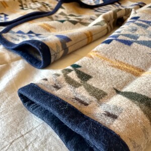 Może przedstawiać: Złożony koc z geometrycznym wzorem w odcieniach beżu, niebieskiego, zieleni i złota. Koc ma ciemnoniebieską obw&oacute;dkę. Wz&oacute;r zawiera tr&oacute;jkąty i kształty przypominające strzały. Koc jest wykonany z miękkiego, teksturowanego materiału.